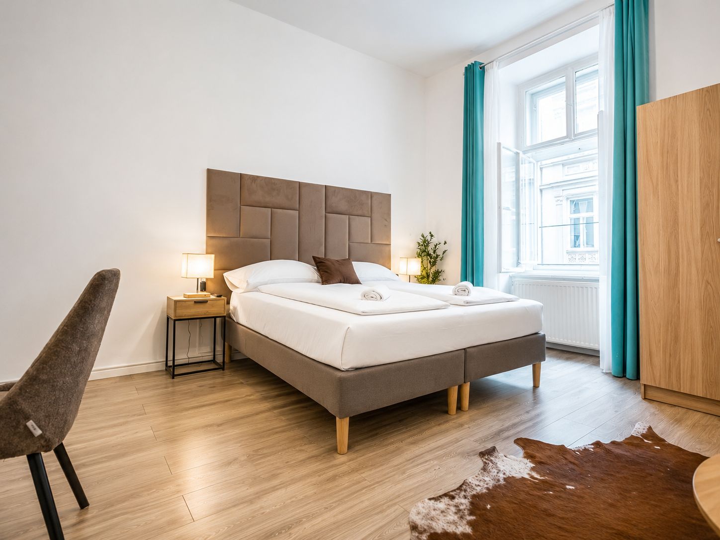 A modern hotel room with a large bed, a nightstand with a lamp, a chair, and a view of the city through a window.