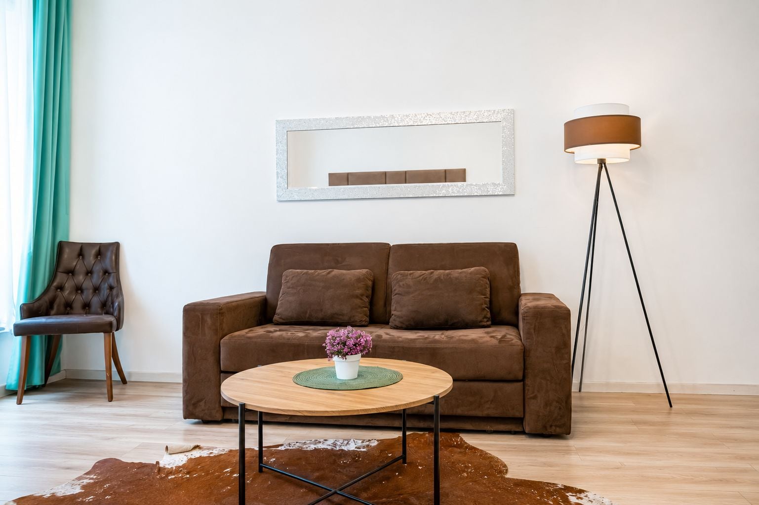 Ein Wohnzimmer mit einem braunen Sofa, einem runden Couchtisch mit Blumenvase, einer Stehlampe und einem Kuhfellteppich.