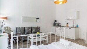 Ett modernt hotellrum med en vit säng, en soffa med mönstrade kuddar, ett matbord, en liten köksdel och ett stort fönster.