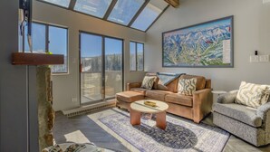 Un salon spacieux avec de grandes fenêtres, un canapé confortable, une table basse et un tableau représentant un paysage de montagne.