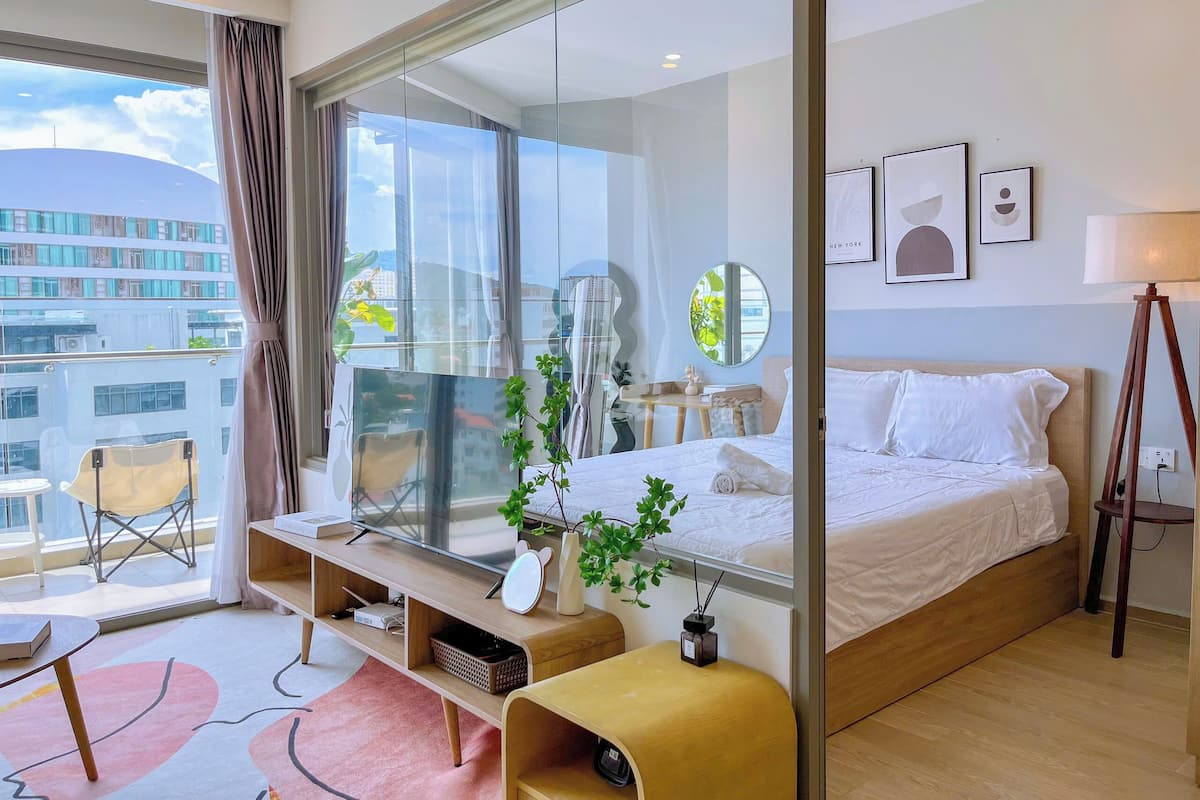 Une chambre d’hôtel moderne dotée d’une grande fenêtre, d’un lit, d’une table de chevet, d’un miroir et d’un petit bureau avec une chaise.
