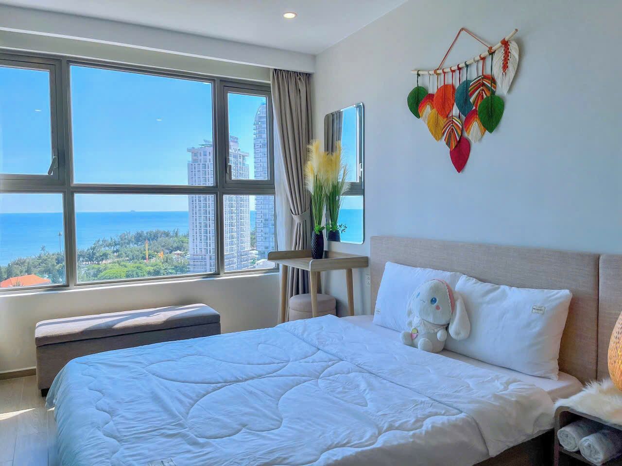 A hotel room with a large bed, a bedside table, a nightstand, a window with a view of the city, and a decorative wall hanging.