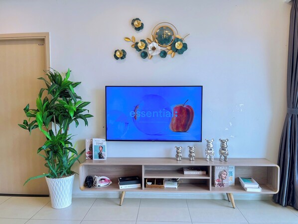 Un salon moderne doté d’un téléviseur à écran plat, d’un élément décoratif mural, d’une plante en pot et d’un meuble de rangement en bois contenant des livres et des objets décoratifs.