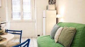 Un ambiente compatto con angolo ristoro, un divano verde con cuscini decorati, un frigorifero bianco e un oggetto decorativo appeso al muro.