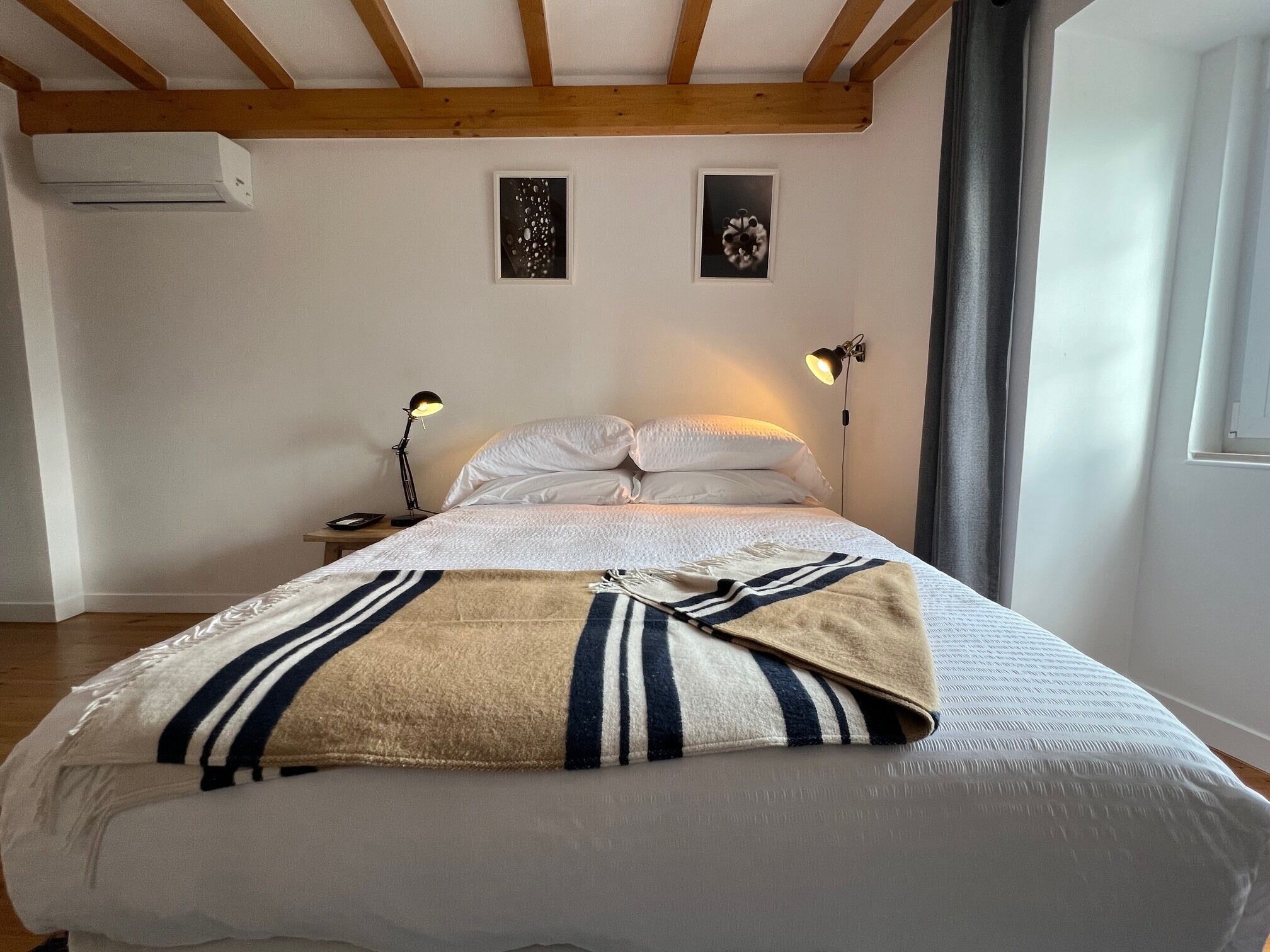 Une chambre à coucher comprenant un lit, deux lampes fixées au mur, une table de chevet, un climatiseur et deux tableaux encadrés accrochés au mur.