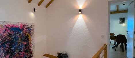 A staircase with wooden beams and a handrail, leading to a room with a painting and a wall-mounted light fixture.