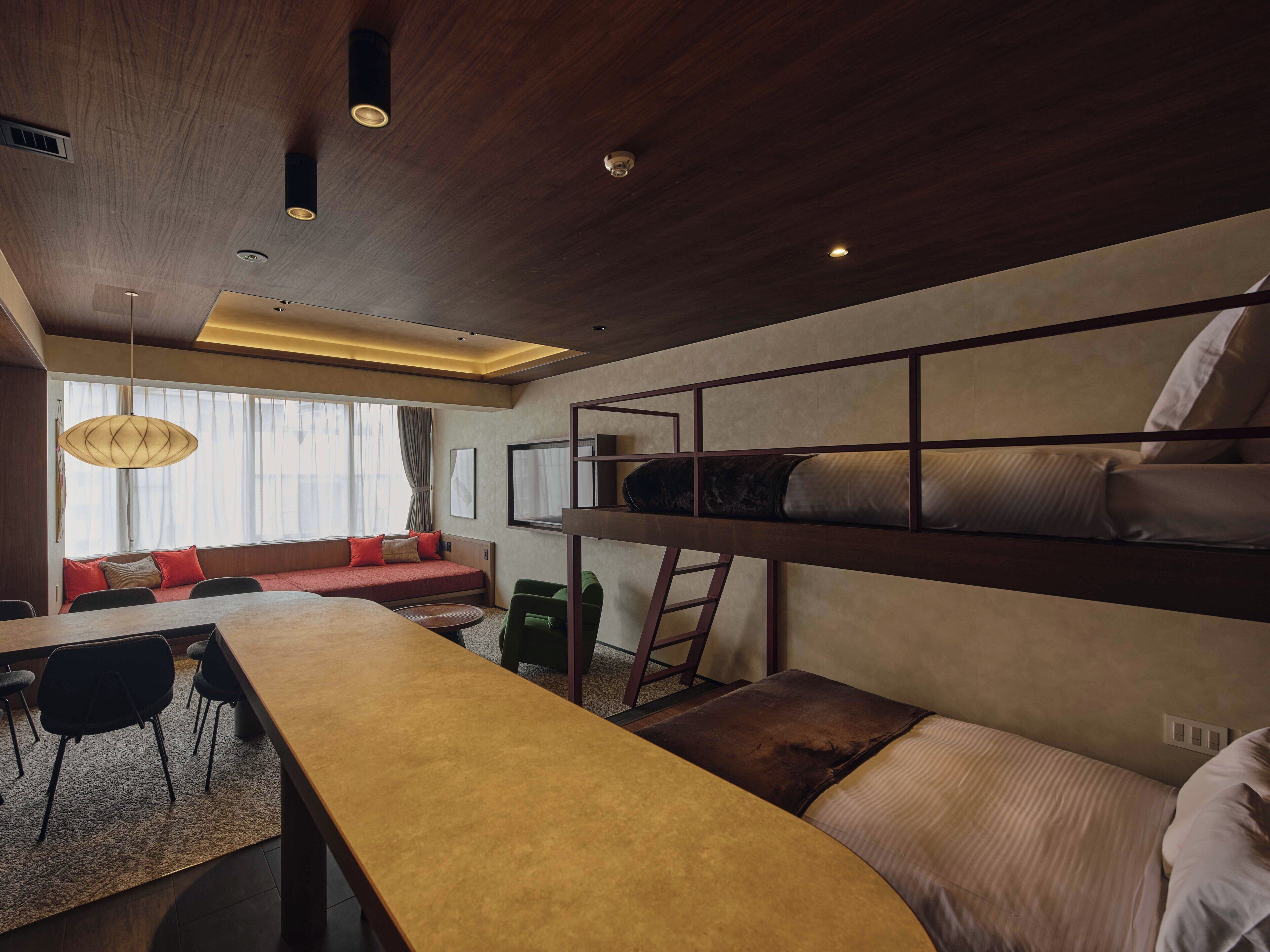 A modern hotel room with a bunk bed, a dining area with a table and chairs, a sofa, and a large window with curtains.