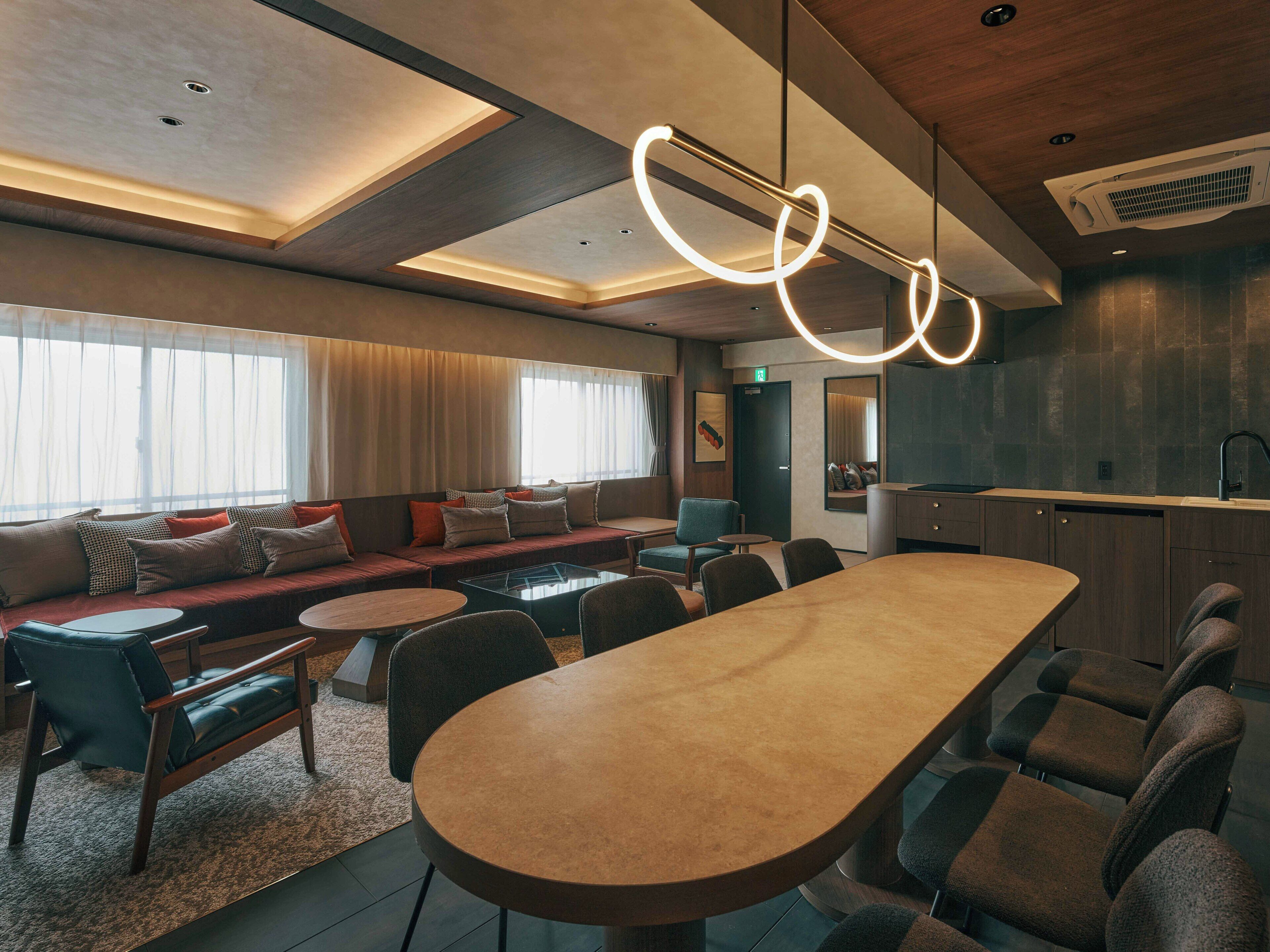 A modern hotel lobby with a long dining table, seating area with sofas, and a kitchen area.