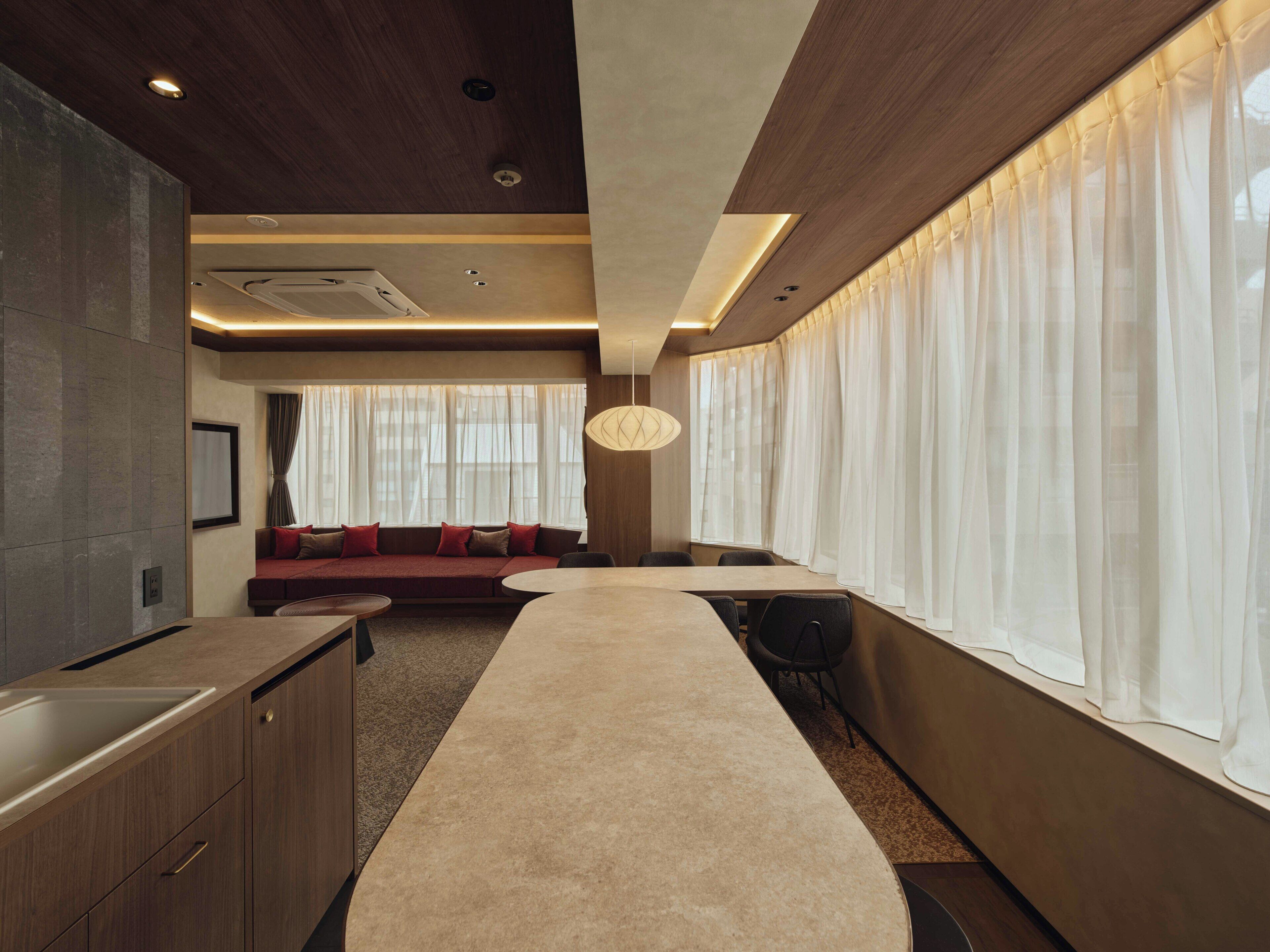 Une chambre d’hôtel moderne avec une longue table à manger, un canapé et une grande fenêtre ornée de rideaux légers.