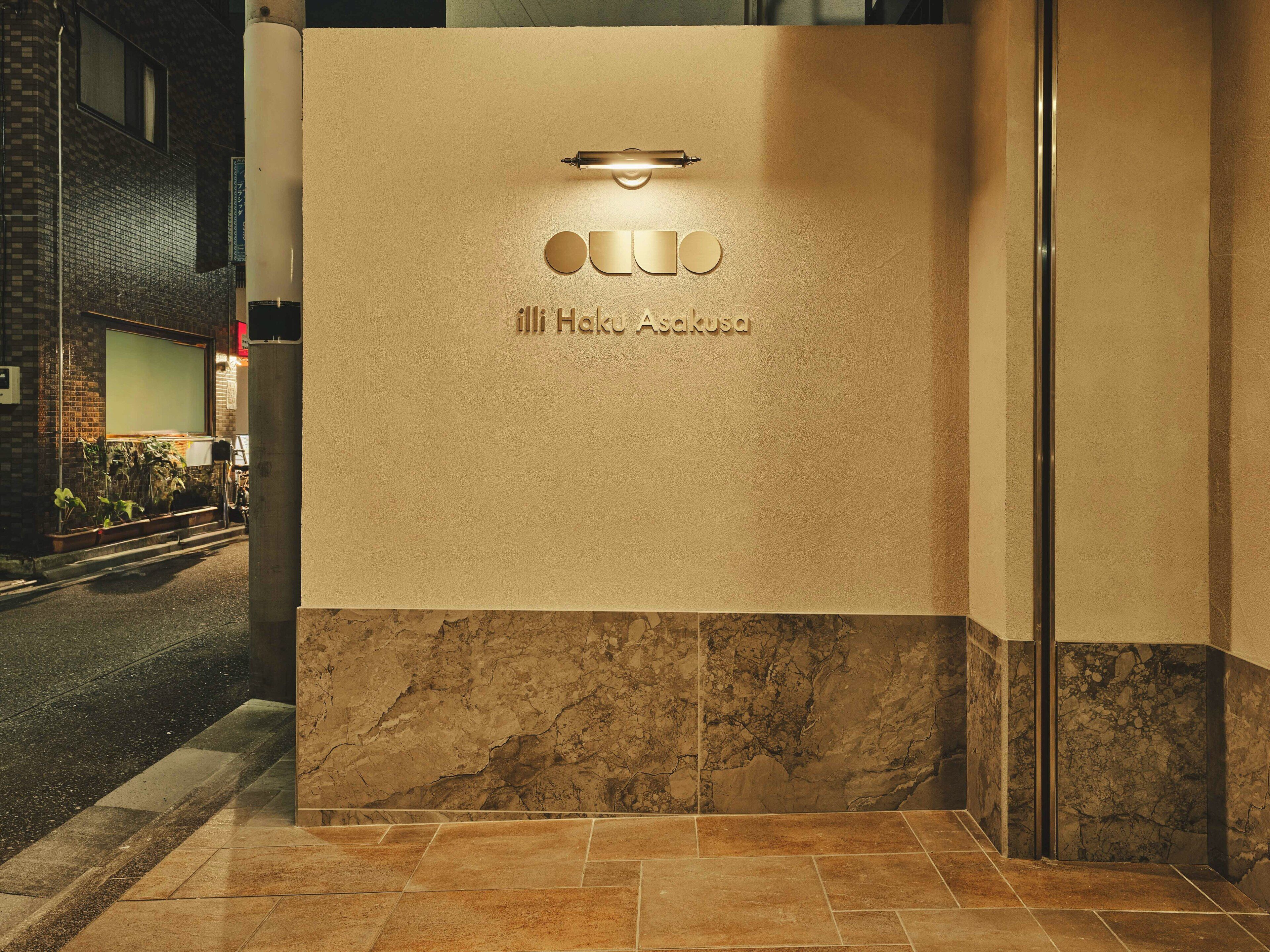 A hotel entrance with a marble floor, a light fixture above the entrance, and the name 'Hako Asakusa' engraved on the wall.