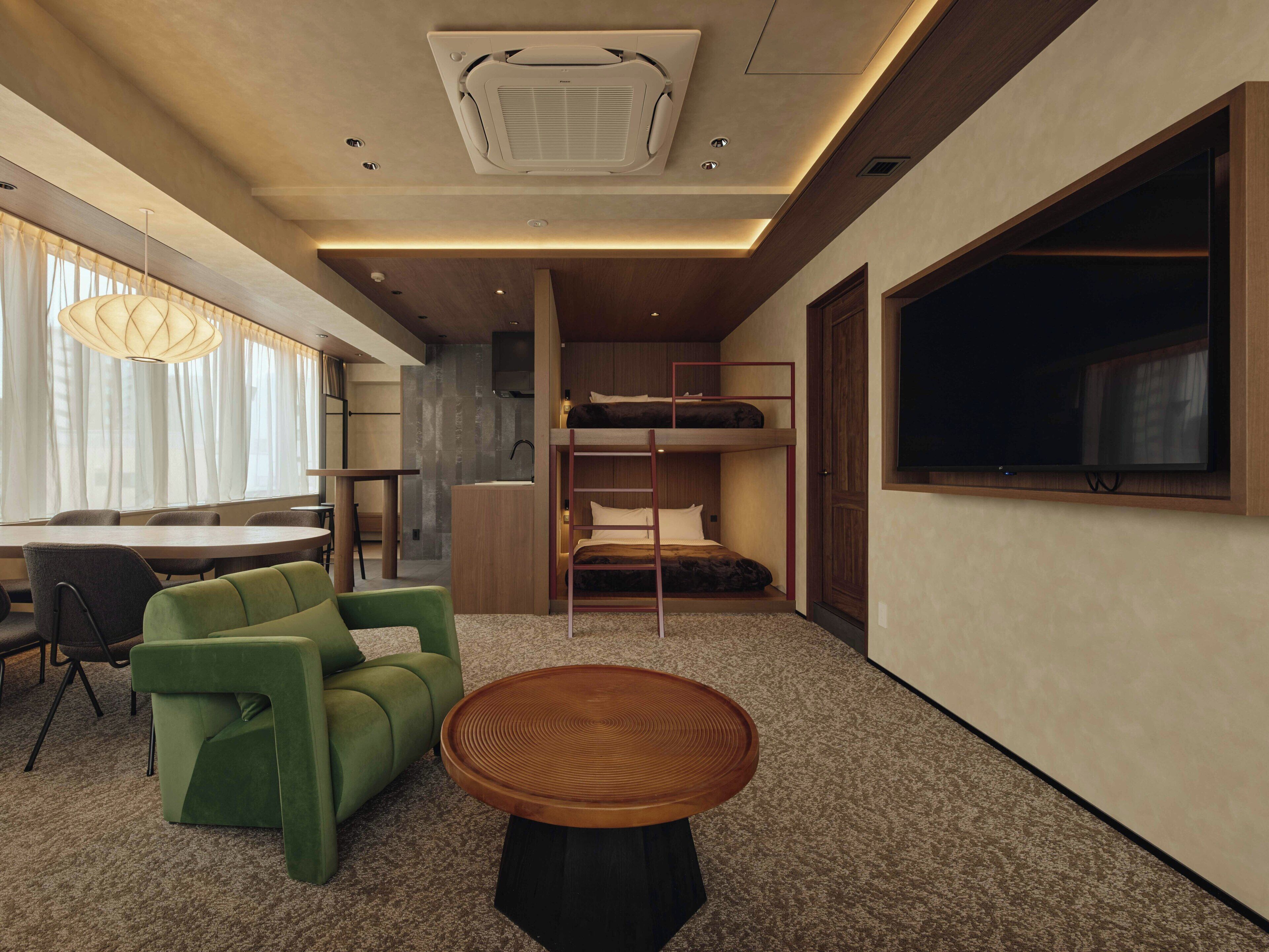 A modern hotel room with a bunk bed, a desk, a green sofa, a round wooden table, and a large window.