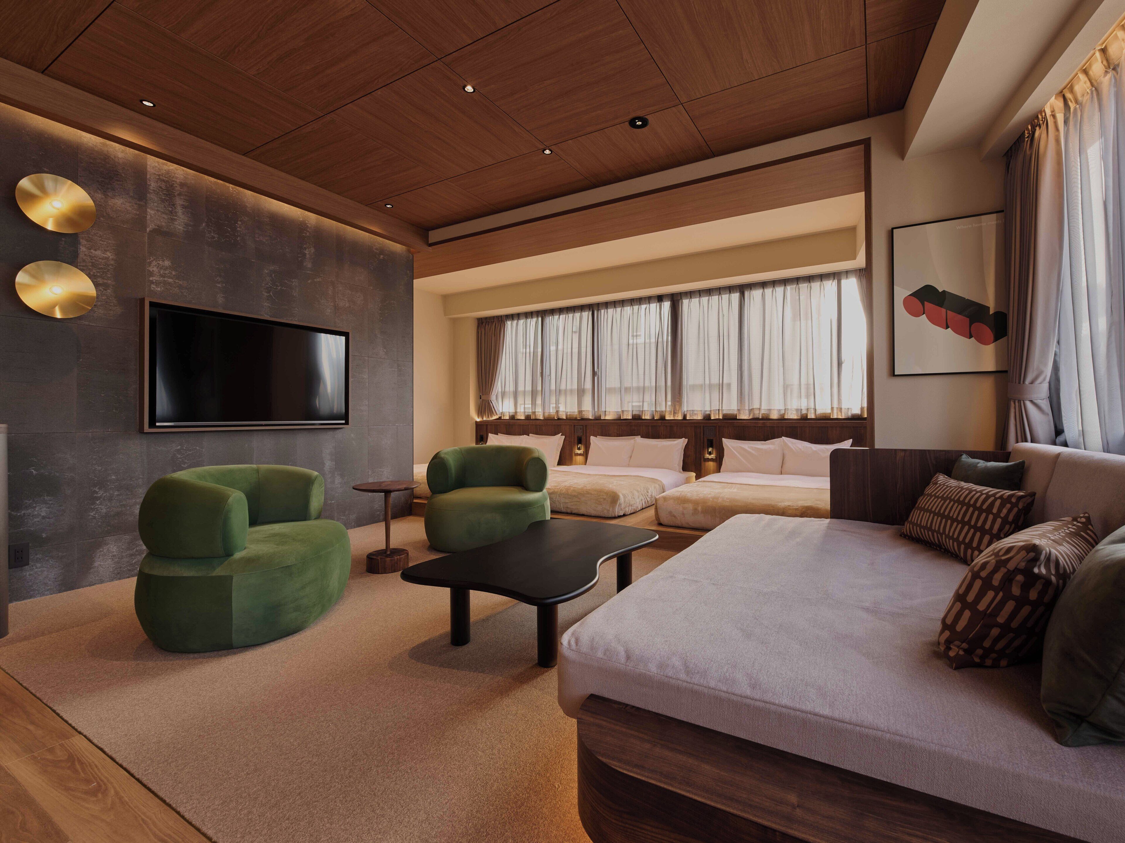 A modern hotel room with a wooden ceiling, a flat-screen TV mounted on the wall, a large sofa, two green armchairs, a small round table, and a window with sheer curtains.