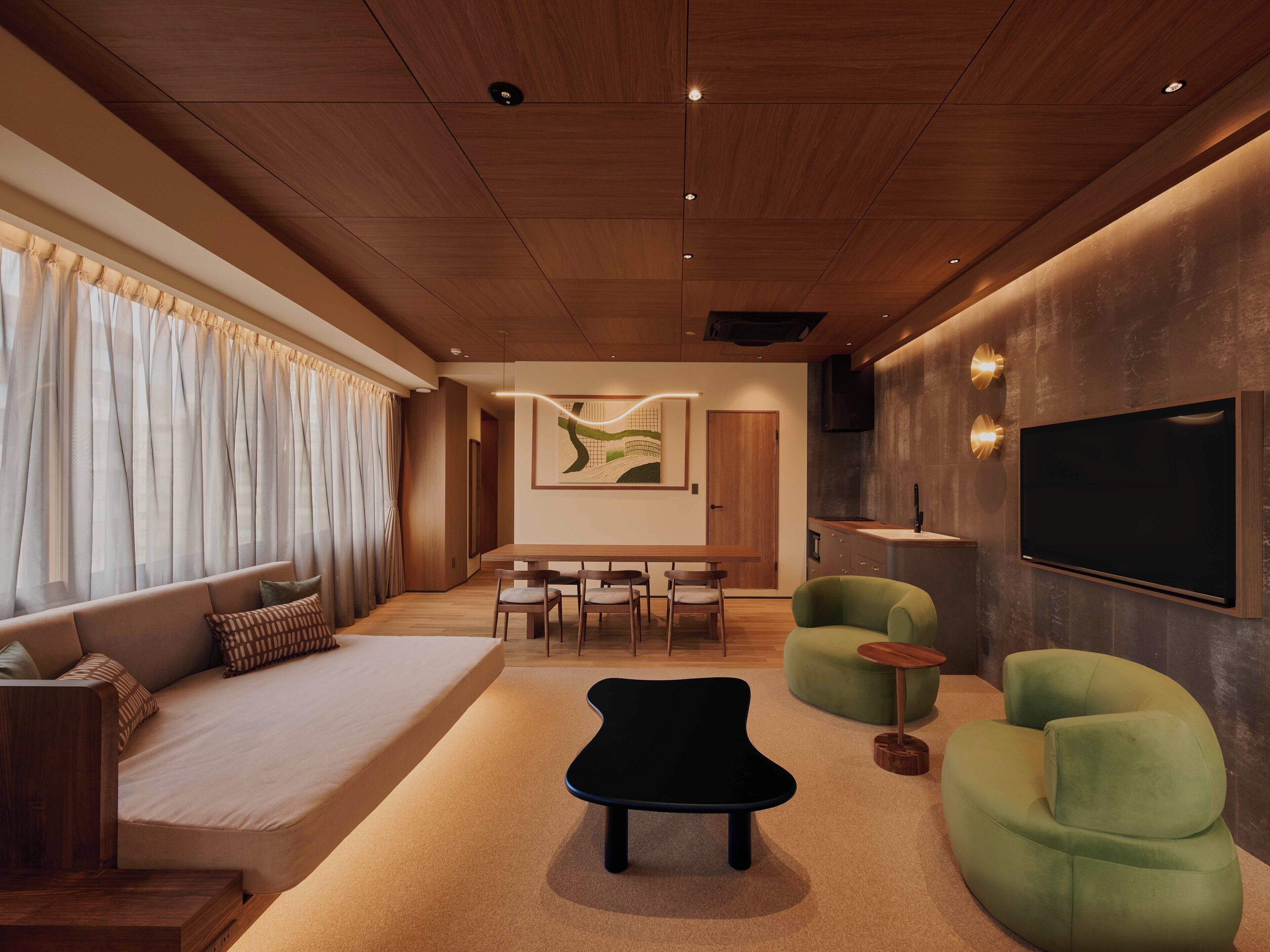 A modern living room with a sofa, a black ottoman, green armchairs, a dining area, and a TV mounted on the wall.