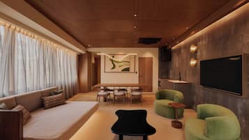 A modern living room with a sofa, a black ottoman, green armchairs, a dining area, and a TV mounted on the wall.