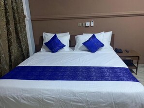A hotel room with a bed, two blue pillows, a white bedspread, and a brown wall. - Sannak Hotel (Sunyani)