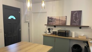 Una cocina moderna con una mesa de madera, un microondas, un tostador y un cubo de basura. Hay dos lámparas colgantes sobre la encimera y una puerta con un ventanal.