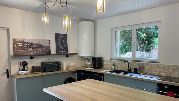 Una cocina moderna con una isla de madera, taburetes de bar, una cafetera, un microondas y un lavavajillas. Hay luces colgantes, un ventanal con vistas a la vegetación y obras de arte en la pared.