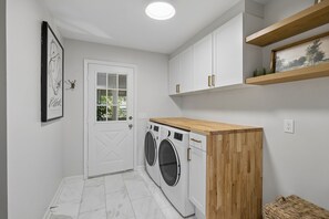 A laundry room with a white door, two front-loading washing machines, a wooden counter, and a shelf with a picture.