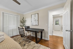 A bedroom with a bed, a desk, a chair, a plant, and a door leading to another room.