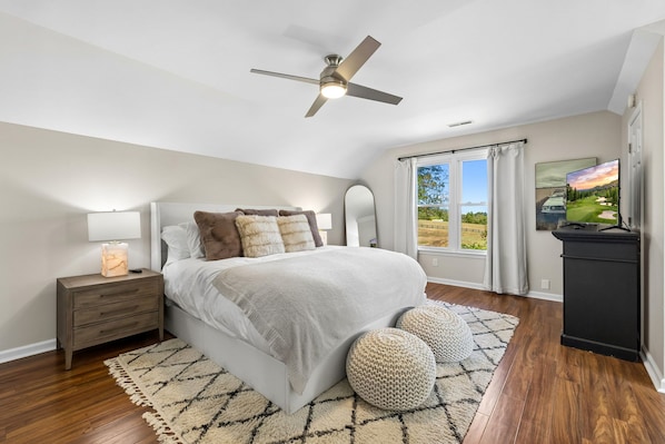 A bedroom with a bed, nightstand, lamp, ceiling fan, large window with curtains, and a TV.