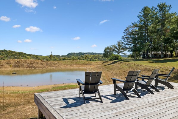 Una terraza de madera con tres sillas Adirondack mirando un lago tranquilo, rodeada de césped y árboles.