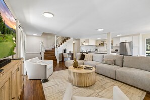 A modern living room with a large flat-screen TV, a staircase, and a kitchen area. - Luxe Franklin Home| 15 Acres w Pond & Game Room (Franklin)
