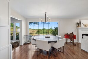 A modern dining area with a round table, white chairs, and a view of a landscape through large windows.