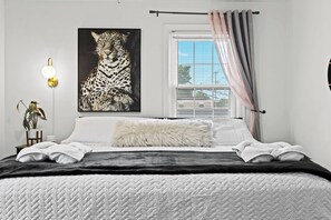 Een slaapkamer met een groot kunstwerk met luipaardprint, een bed met wit en zwart beddengoed, een zacht kussen, een nachtkastje met een lamp en een raam met gordijnen.