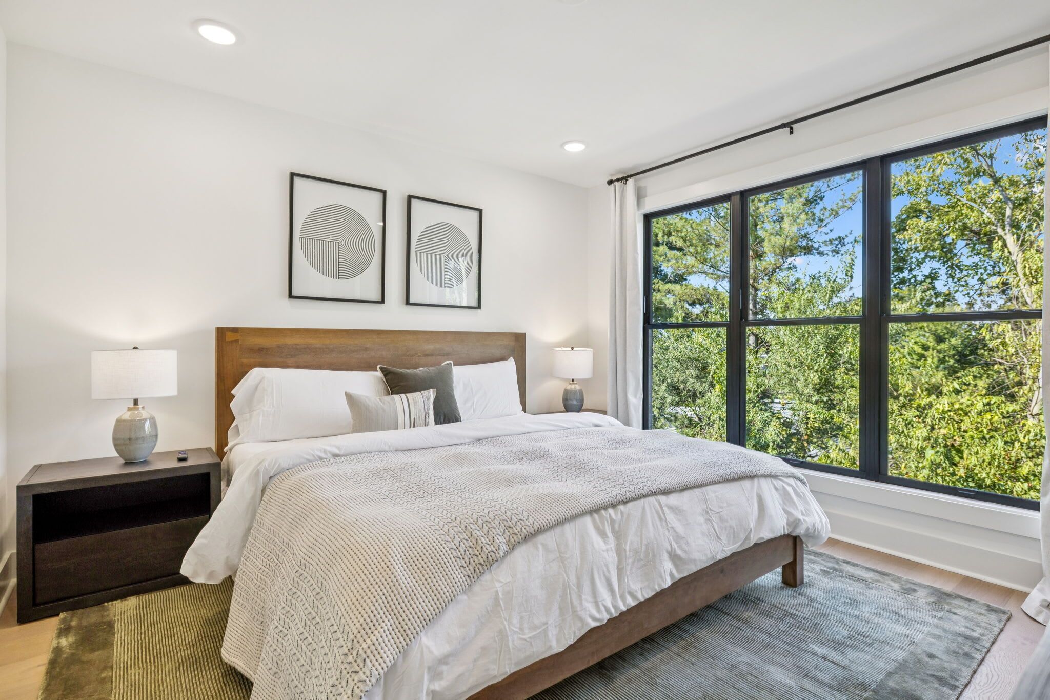 A modern bedroom with a large bed, two bedside tables with lamps, a headboard, and a large window with a view of trees.