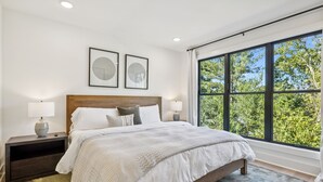 A modern bedroom with a large bed, two bedside tables with lamps, a headboard, and a large window with a view of trees. - Luxury retreat with exclusive rooftop hot tub (Nashville)
