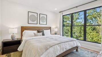 A modern bedroom with a large bed, two bedside tables with lamps, a headboard, and a large window with a view of trees.