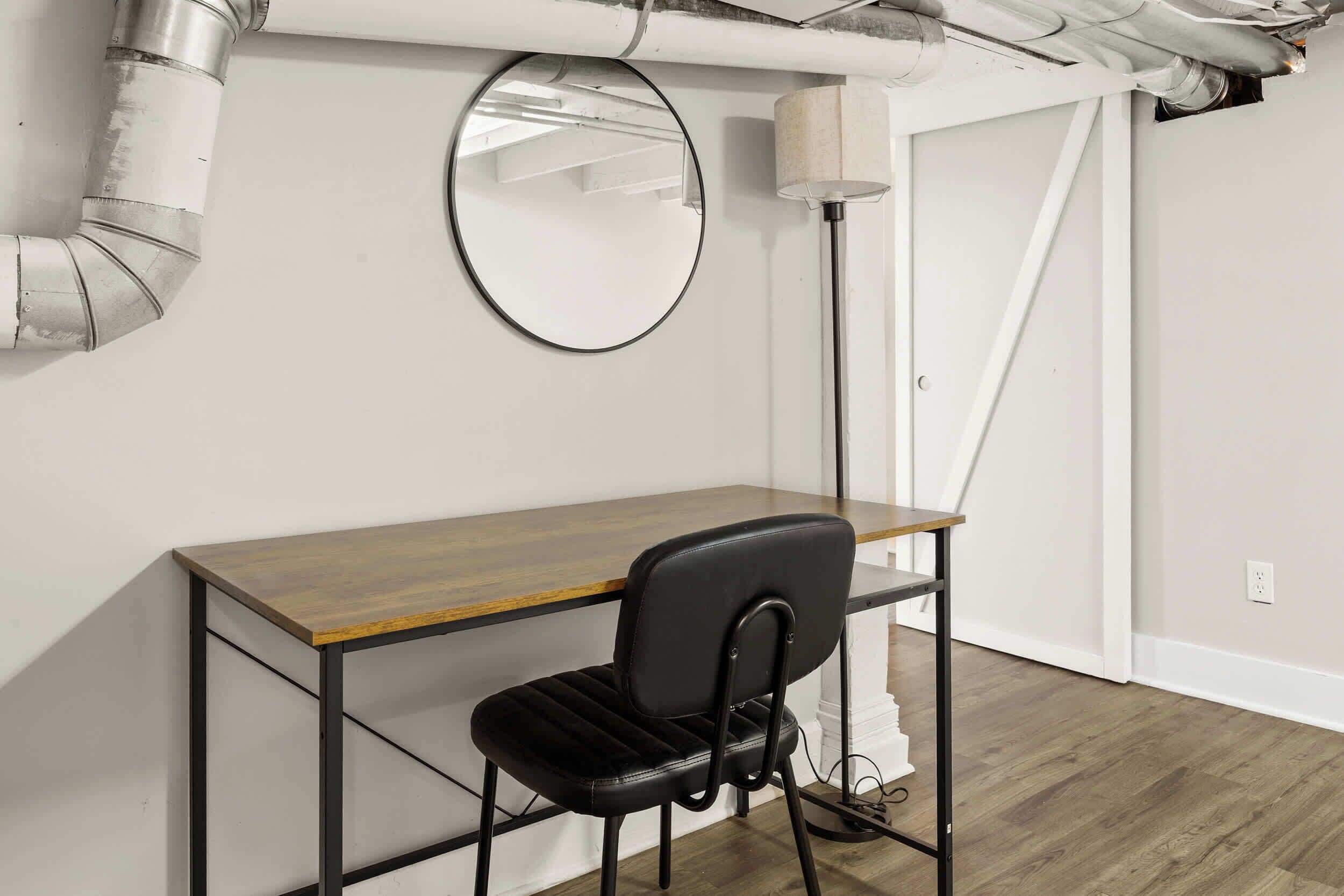 Un espacio de trabajo con un escritorio de madera, una silla negra, un espejo redondo y una lámpara de pie.