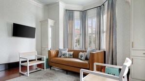 Una sala de estar con un sofá marrón, sillas blancas, un televisor de pantalla plana y cortinas estampadas.