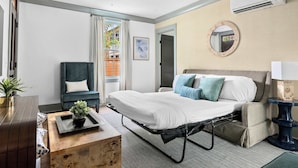 Una habitación de hotel moderna con una cama, una silla, una mesita con una planta y un equipo de aire acondicionado.