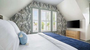 Een slaapkamer met schuine balken, een bed met witte beddengoed en blauwe accenten, een houten dressoir en een aan de muur bevestigde televisie.