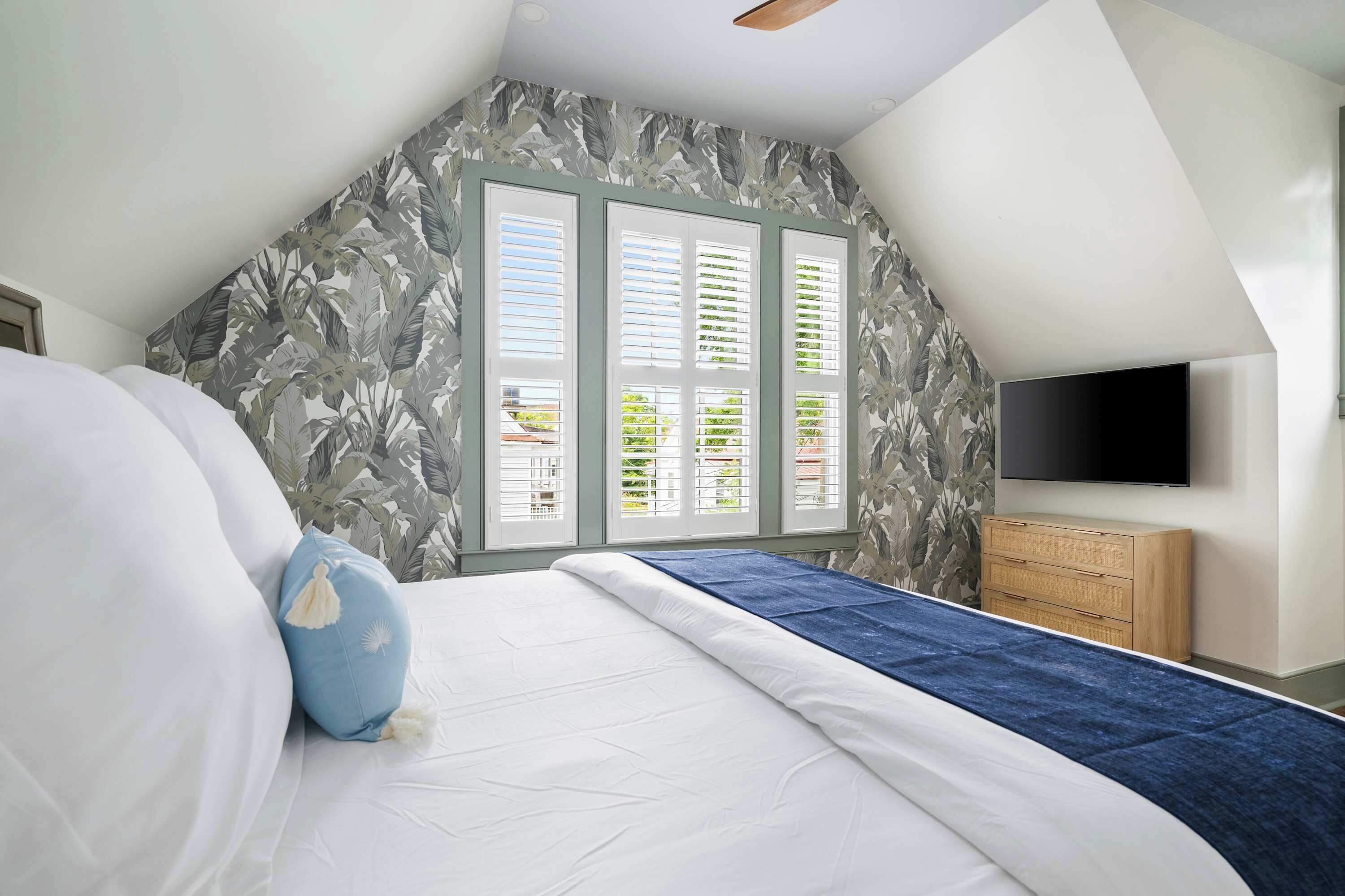 Quarto com teto inclinado, cama com roupa de cama branca e detalhes azuis, cómoda de madeira e televisão fixada na parede.