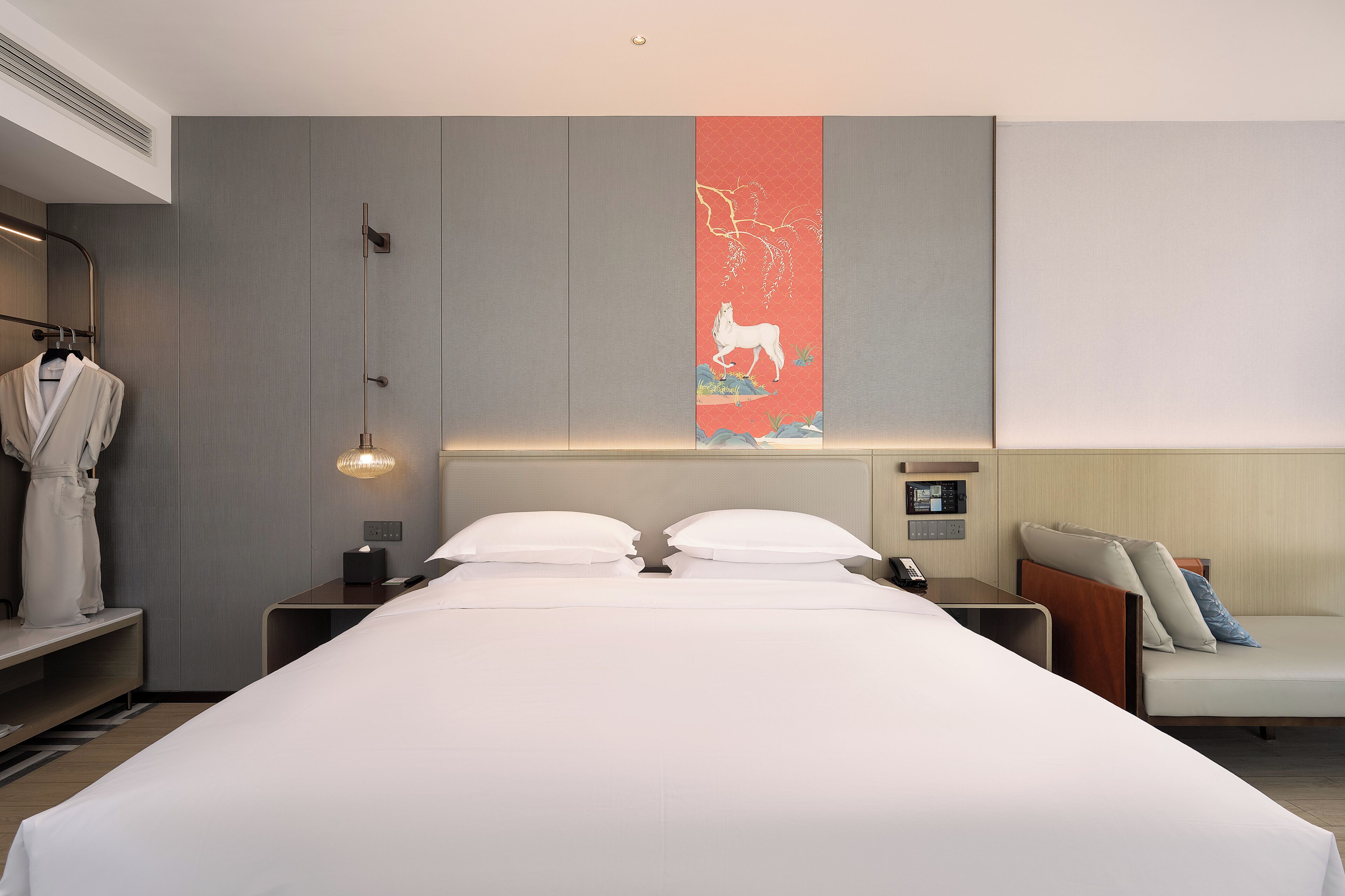 Een moderne hotelkamer met een groot bed, een nachtkastje met een lamp, een badjas hangend aan een rek, en een kunstwerk aan de muur.