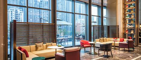 A spacious lobby with a high ceiling, large windows offering a view of tall buildings, a seating area with sofas and chairs, and a bookshelf filled with books.