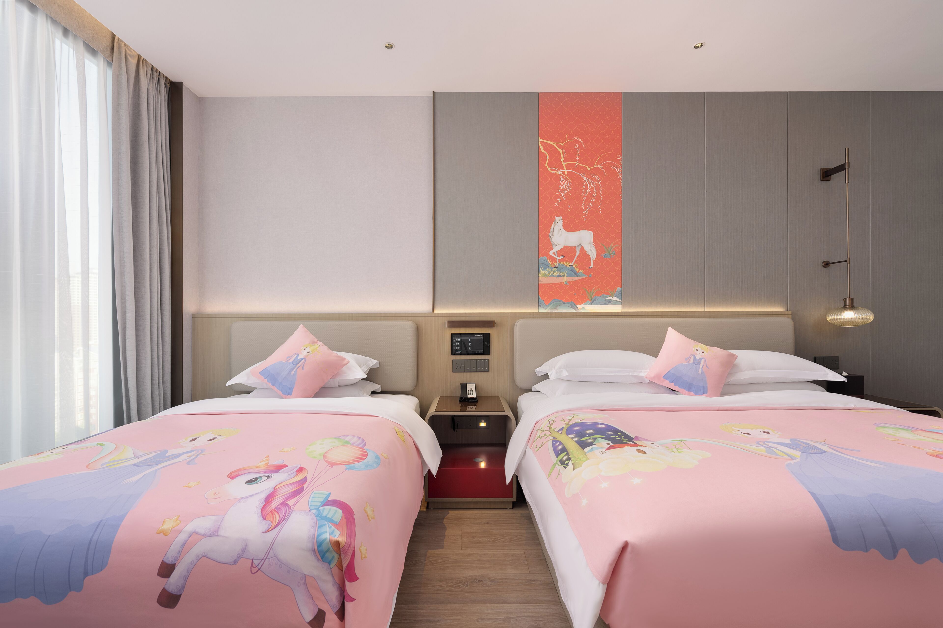 A hotel room with two beds, each with a pink unicorn-themed bedspread, a nightstand with a phone, and a wall-mounted light fixture.