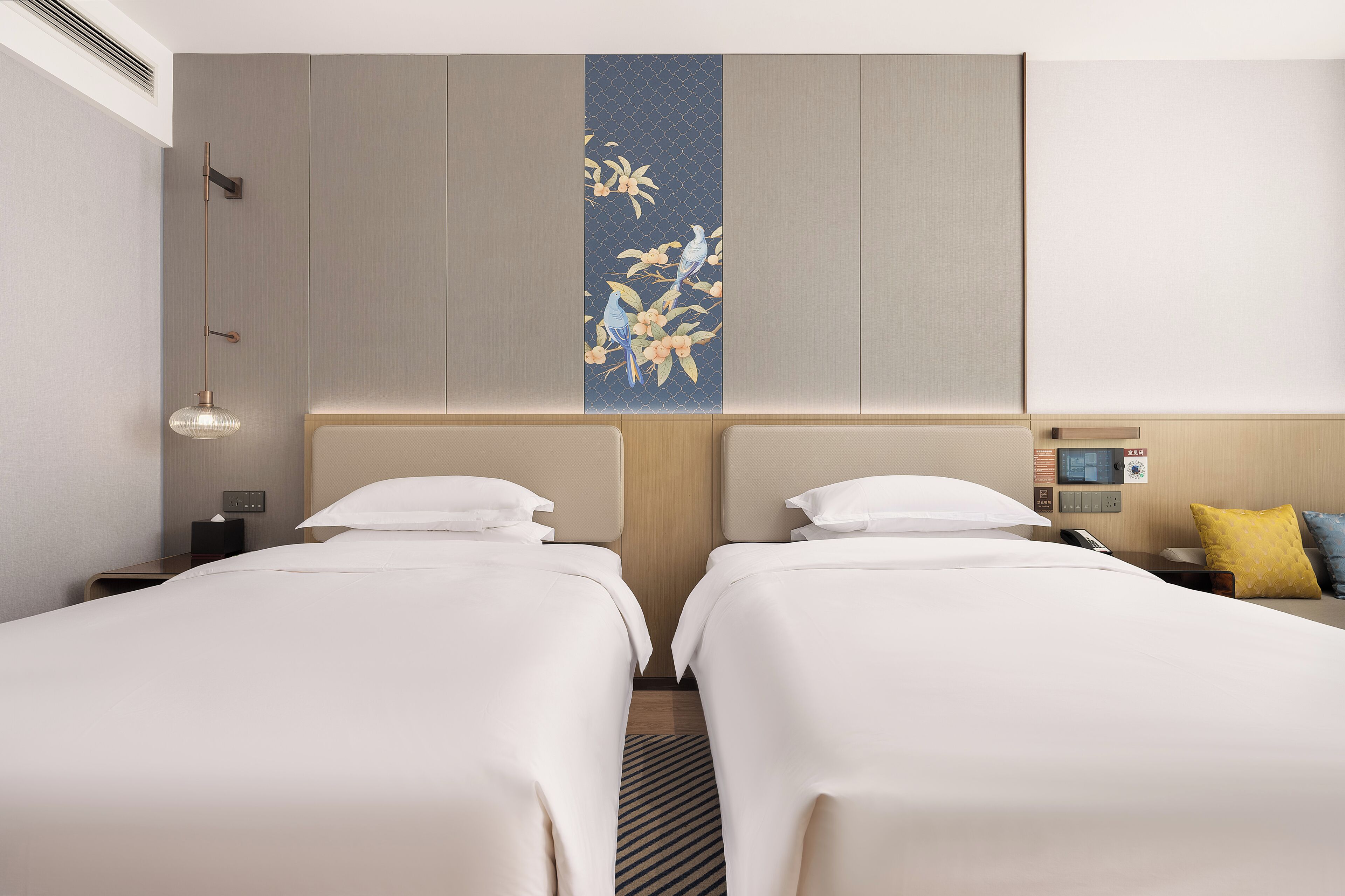 A hotel room with two beds, a wall-mounted TV, a bedside table with a lamp, and a decorative wall panel.