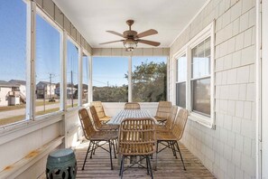 Varanda coberta com mesa, cadeiras, ventilador de teto e grandes janelas.