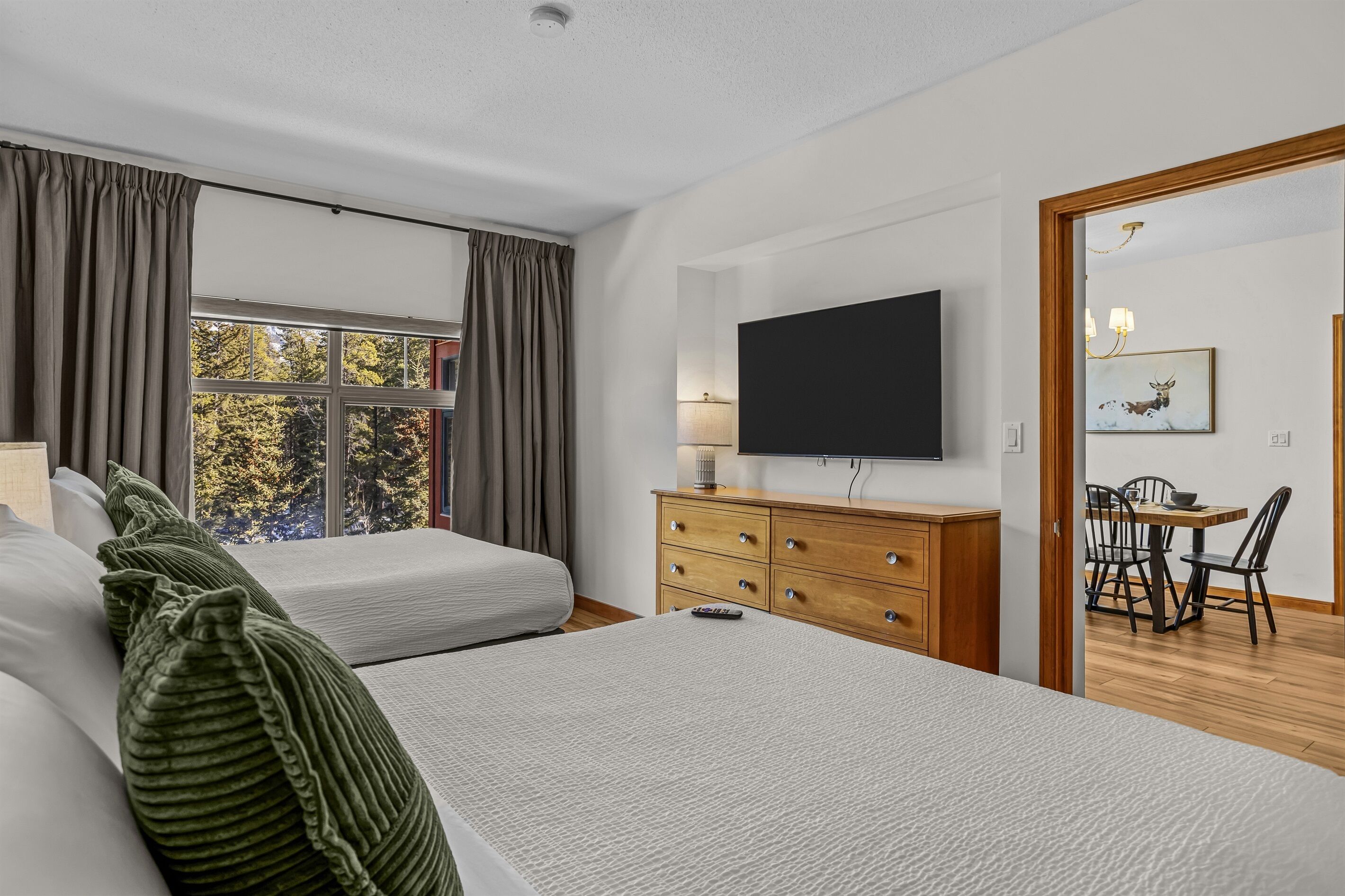 A modern hotel room with two beds, a flat-screen TV, a wooden dresser, and a dining area with a table and chairs.