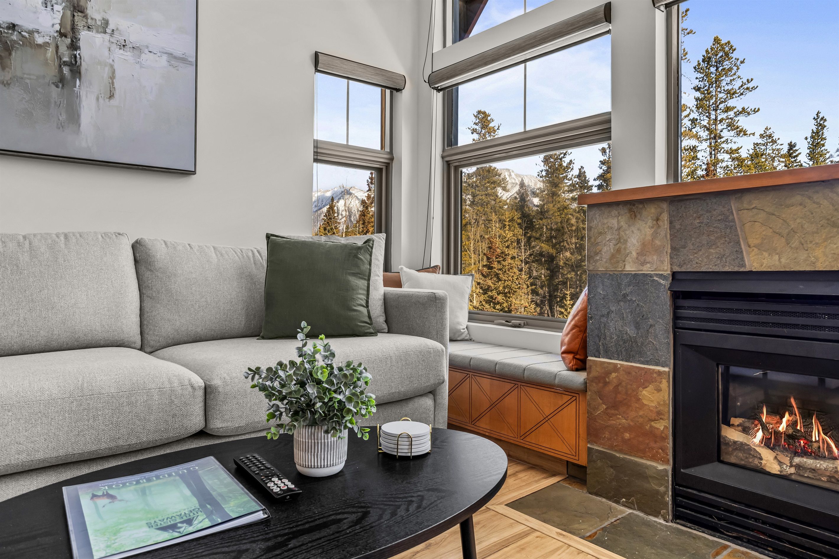 A modern living room with a grey sofa, a fireplace, and large windows offering a view of trees and mountains.