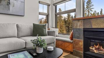 A modern living room with a grey sofa, a fireplace, and large windows offering a view of trees and mountains.