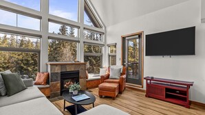 Un salon spacieux doté d’une cheminée, de fauteuils en cuir, d’un téléviseur à écran plat et de grandes fenêtres offrant une vue sur les arbres.