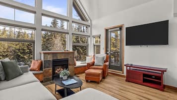 A spacious living room with a fireplace, leather seating, a flat-screen TV, and large windows offering a view of trees.
