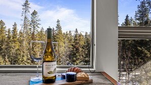 Une bouteille de vin, un verre, un croissant et un morceau de fromage, disposés sur une planche en bois près d’une fenêtre donnant sur une forêt.