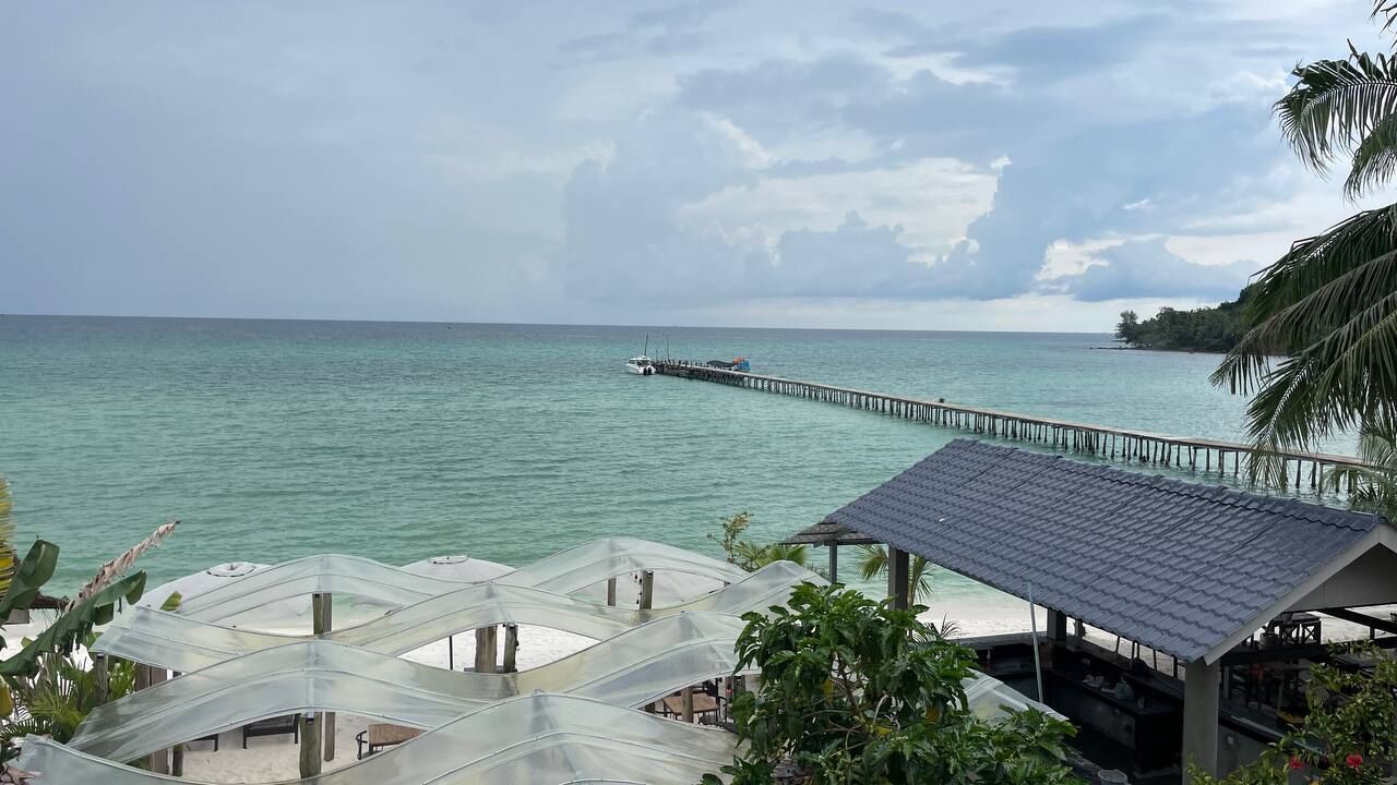 A beachside view with a pier extending into the ocean, a covered seating area with a roof, and lush tropical vegetation.