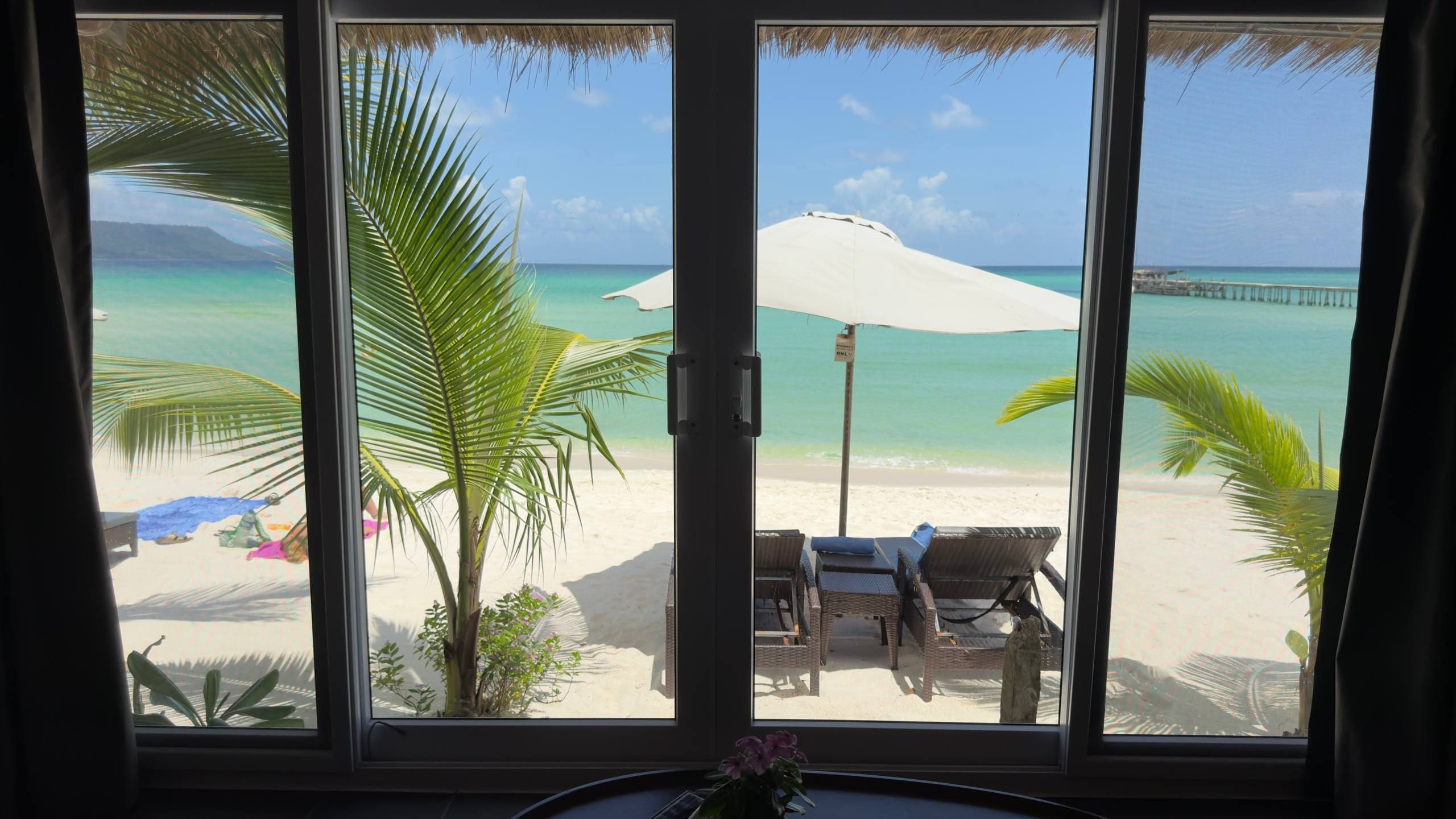 A beach view through a window with a white umbrella, lounge chairs, and a clear sky.