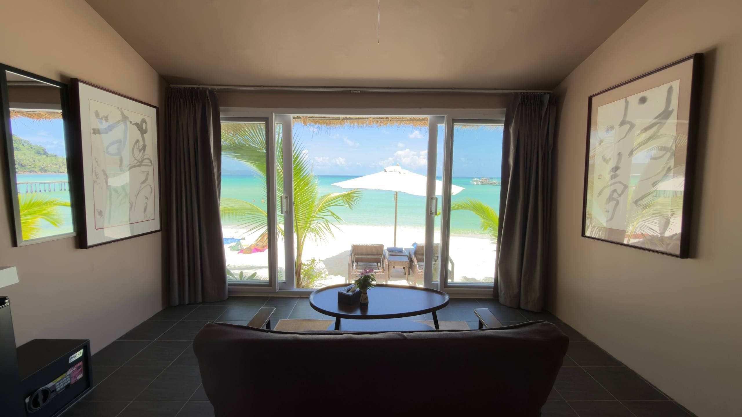 A hotel room with a large sliding glass door leading to a beach view, a round table, and two framed artworks on the walls.